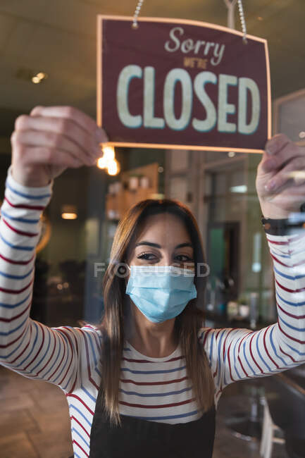 Coiffeuse caucasienne travaillant dans un salon de coiffure portant un masque facial, tenant Désolé, nous sommes fermés signe. Santé et hygiène sur le lieu de travail pendant la pandémie de Coronavirus Covid 19. — Photo de stock