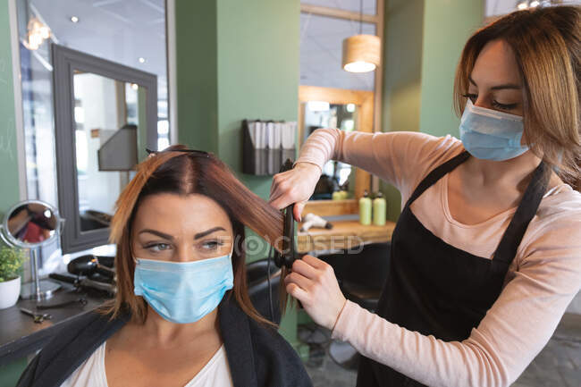 Белая женщина-парикмахер работает в парикмахерской в маске для лица, выпрямляя волосы белой клиентки в маске. Здоровье и гиперактивность на рабочем месте во время коронавируса Ковид 19 пандемии. — стоковое фото