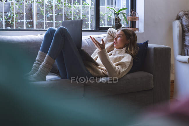 Mujer caucásica pasar tiempo en casa, acostado en el sofá en la sala de estar con ordenador portátil para videollamada. Distanciamiento social durante el bloqueo de cuarentena del Coronavirus Covid 19. - foto de stock