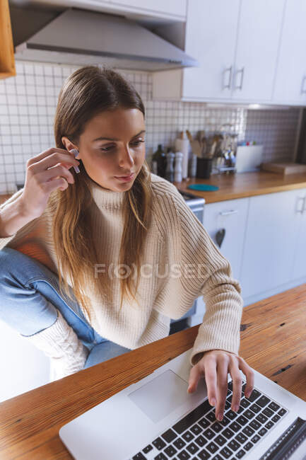 Caucasian woman spending time at home, sitting in kitchen using laptop computer, putting her earphones on. Social distancing during Covid 19 Coronavirus quarantine lockdown. — Stock Photo