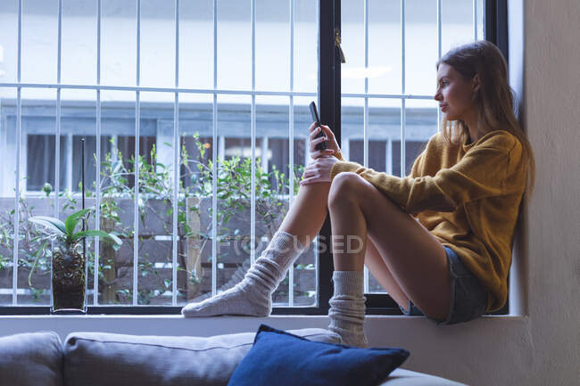 Mulher caucasiana passar o tempo em casa, sentado no peitoril da janela na sala de estar, usando smartphone. Distanciamento social durante o bloqueio de quarentena do Covid 19 Coronavirus. — Fotografia de Stock
