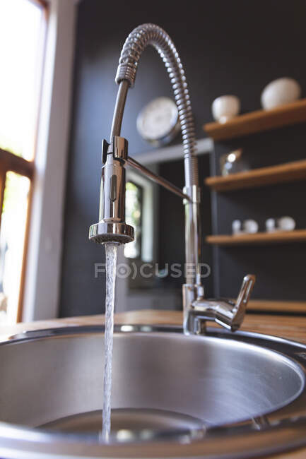 Primo piano di un rubinetto da cucina con acqua che scorre in un lavandino in acciaio in una cucina moderna con ripiani sfocati sullo sfondo. Interni design idea cucina. — Foto stock