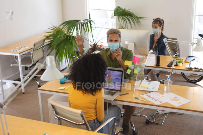 Grupo multiétnico de creativos masculinos y femeninos que trabajan en escritorios de oficina con pantallas protectoras, utilizando computadoras. Salud e higiene en el lugar de trabajo durante la pandemia de Coronavirus Covid 19. - foto de stock