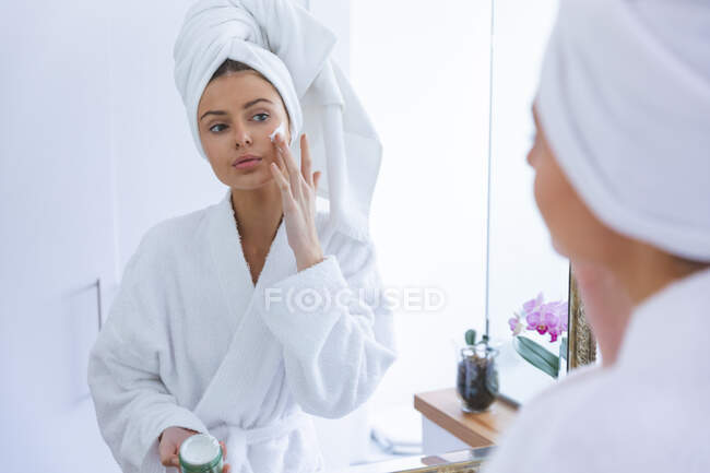 Caucasian woman spending time at home, standing in bathroom, looking in mirror applying face cream. Social distancing during Covid 19 Coronavirus quarantine lockdown. — Stock Photo
