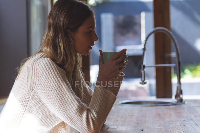 Caucasian woman spending time at home, sitting in kitchen with earphones on, holding green mug. Social distancing during Covid 19 Coronavirus quarantine lockdown. — Stock Photo