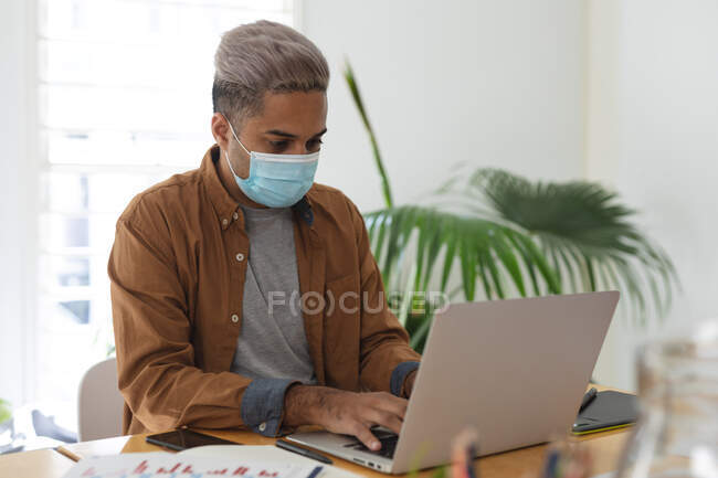 Mixed race male creative business working in modern office wearing face mask. Health and hygiene in the workplace during Coronavirus Covid 19 pandemic. — Stock Photo