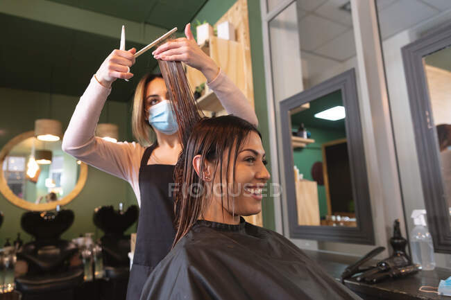 Белая женщина-парикмахер работает в парикмахерской в маске, стрижет волосы белой женщины-клиента. Здоровье и гиперактивность на рабочем месте во время коронавируса Ковид 19 пандемии. — стоковое фото