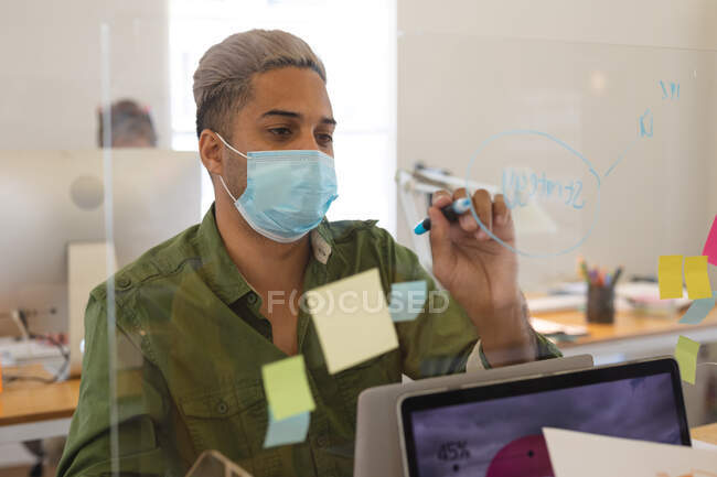 Masculino de raza mixta creativo en mascarilla que trabaja en el escritorio en la oficina, escribiendo en la pantalla protectora. Salud e higiene en el lugar de trabajo durante la pandemia de Coronavirus Covid 19. - foto de stock