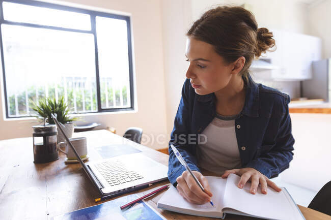 Caucasian woman spending time at home, sitting by table in kitchen using laptop computer, working from home, making notes. Social distancing during Covid 19 Coronavirus quarantine lockdown. — Stock Photo