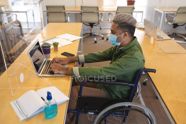 Змішана раса чоловіча творчість сидить на інвалідному візку за столом в офісі, одягнена в маску обличчя, за допомогою ноутбука. Здоров 