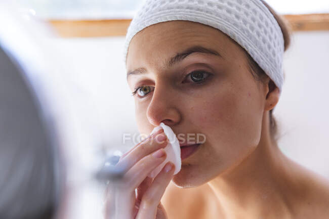 Белая женщина проводит время дома, в ванной, глядя в зеркало, очищая лицо хлопковой салфеткой. Социальное дистанцирование во время изоляции коронавируса Covid 19. — стоковое фото