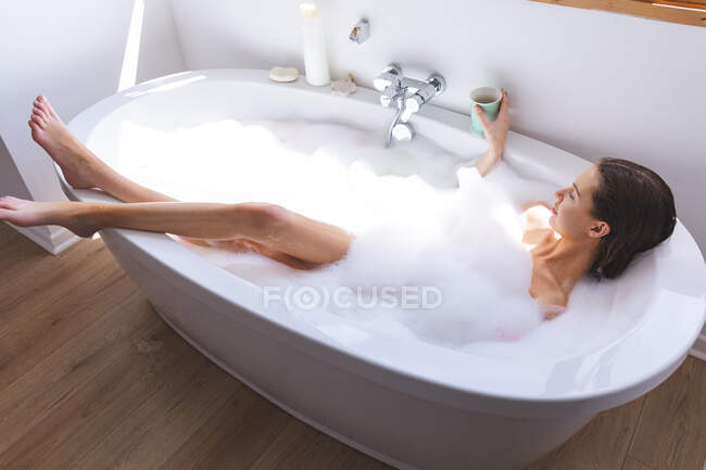 Кавказка проводит время дома, в ванной, лежит в ванне, расслабляясь, держа чашку. Социальное дистанцирование во время изоляции коронавируса Covid 19. — стоковое фото