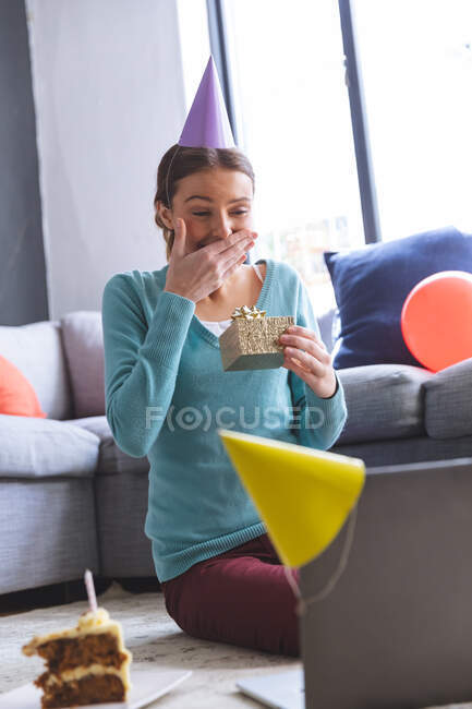 Счастливая кавказская женщина проводит время дома, в шляпе, сидя на полу с помощью компьютера во время видеочата, держа в руках подарок. Социальное дистанцирование во время изоляции коронавируса Covid 19. — стоковое фото