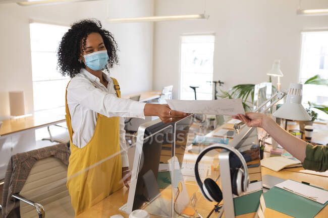 Mixed race female creative working at desks, woman passing document over screen, wearing face mask. Health and hygiene in workplace during Coronavirus Covid 19 pandemic. — Stock Photo