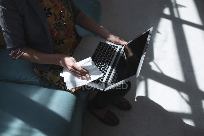 Female business creative sitting on sofa in an office disinfecting laptop computer with a wipe. Health and hygiene in workplace during Coronavirus Covid 19 pandemic. — Stock Photo