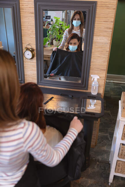 Kaukasische Friseurin, die im Friseursalon arbeitet, trägt Gesichtsmaske und setzt einer kaukasischen Kundin einen Umhang auf. Gesundheit und Hygiene am Arbeitsplatz während der Coronavirus Covid 19 Pandemie. — Stockfoto