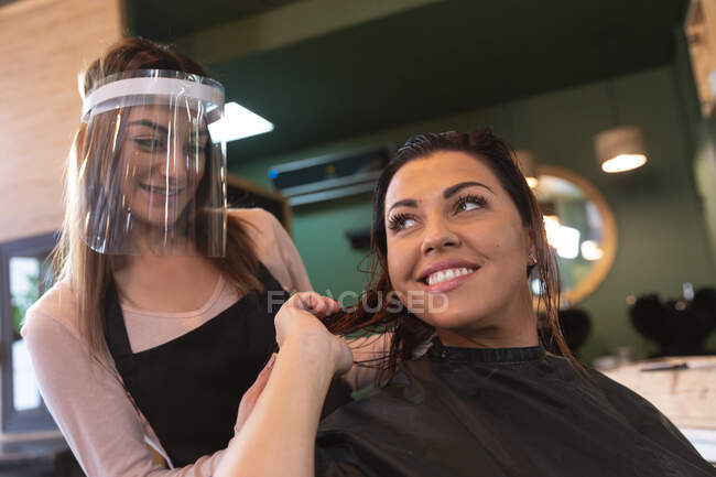 Белая женщина-парикмахер, работающая в парикмахерской в маске, расчесывающая волосы кавказской клиентки, улыбающейся. Здоровье и гиперактивность на рабочем месте во время коронавируса Ковид 19 пандемии. — стоковое фото