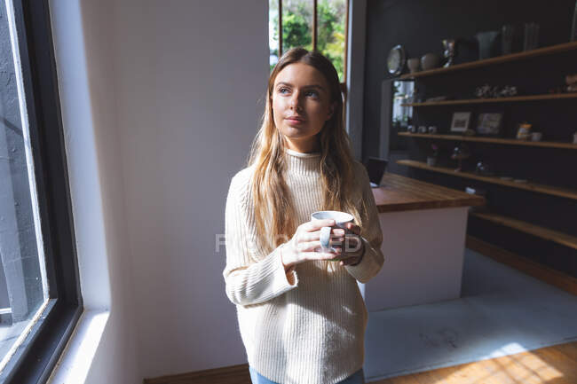 Caucasian woman spending time at home, standing in kitchen, holding green mug looking out of window. Social distancing during Covid 19 Coronavirus quarantine lockdown. — Stock Photo