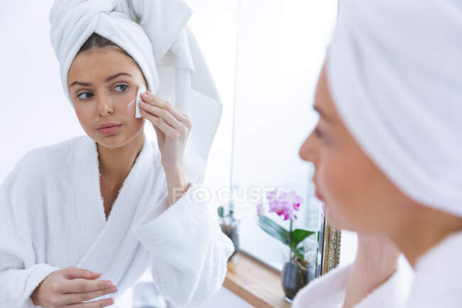 Caucasian woman spending time at home, standing in bathroom, looking in mirror removing make up with cotton pad. Social distancing during Covid 19 Coronavirus quarantine lockdown. — Stock Photo
