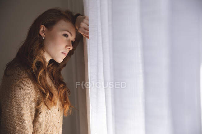 An attractive, ginger Caucasian woman spending time at home, in living room, looking out of the window. Social distancing during Covid 19 Coronavirus quarantine lockdown. — Stock Photo