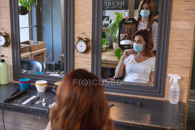 Белая женщина-парикмахер работает в парикмахерской в маске, показывая стрижку белой женщине в маске. Здоровье и гиперактивность на рабочем месте во время коронавируса Ковид 19 пандемии. — стоковое фото