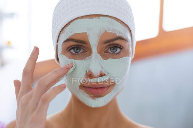 Портрет белой женщины, проводящей время дома, стоящей в ванной, смотрящей в камеру в маске для лица. Социальное дистанцирование во время изоляции коронавируса Covid 19. — стоковое фото