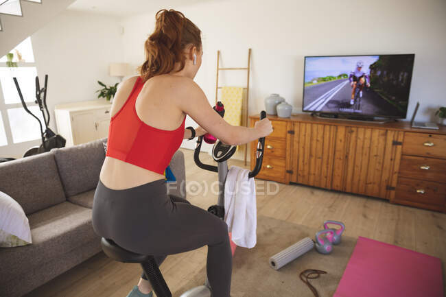 Белая женщина проводит время дома, в гостиной, тренируется на стационарном велосипеде, смотрит телевизор. Социальное дистанцирование во время изоляции коронавируса Covid 19. — стоковое фото