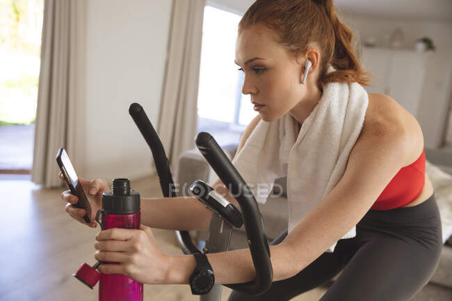 Mujer caucásica pasando tiempo en casa, en la sala de estar, haciendo ejercicio en bicicleta estacionaria, usando su teléfono inteligente. Distanciamiento social durante el bloqueo de cuarentena del Coronavirus Covid 19. - foto de stock