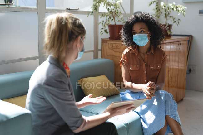 Mixed race and Caucasian female business creatives wearing face masks and distancing on sofa, talking and using tablet in office. Health and hygiene in workplace during Coronavirus Covid 19 pandemic. — Stock Photo
