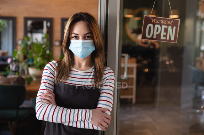 Caucasian female hairdresser working in hair salon wearing face mask, leaning on glass door with sign saying Yes, We Are Open. Health and hygiene in workplace during Coronavirus Covid 19 pandemic. — Stock Photo