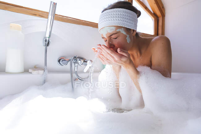 Mujer caucásica pasar tiempo en casa, en el baño, sentado en la bañera, enjuagar la máscara de la cara. Distanciamiento social durante el bloqueo de cuarentena del Coronavirus Covid 19. - foto de stock