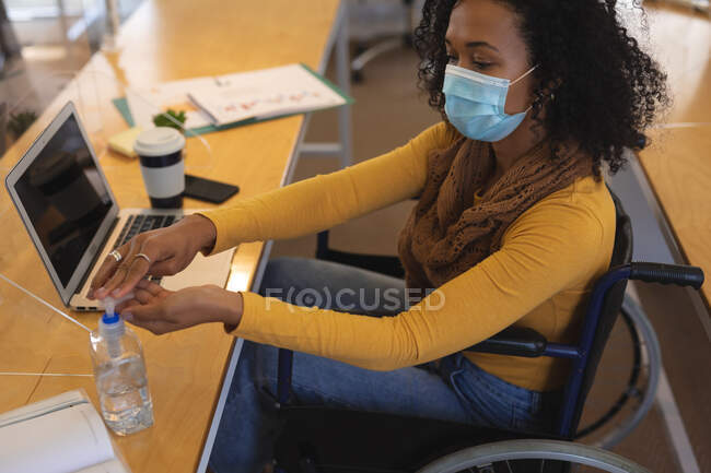 Creativa mista di razza femminile seduta su una sedia a rotelle alla scrivania in un ufficio, indossando una maschera facciale, disinfettando le mani con antipasto per le mani. Salute e igiene sul luogo di lavoro durante la pandemia di Coronavirus Covid 19. — Foto stock