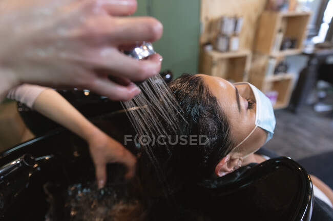 Peluquería femenina caucásica que trabaja en peluquería con mascarilla facial, lavado de cabello de cliente caucásico femenino en mascarilla facial. Salud e higiene en el lugar de trabajo durante la pandemia de Coronavirus Covid 19. - foto de stock