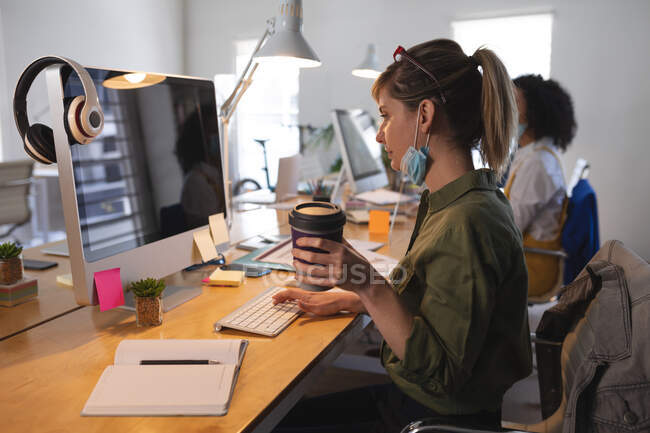 Femme caucasienne portant un masque facial abaissé assis au bureau avec des collègues utilisant un ordinateur et tenant du café. Santé et hygiène sur le lieu de travail pendant la pandémie de Coronavirus Covid 19. — Photo de stock