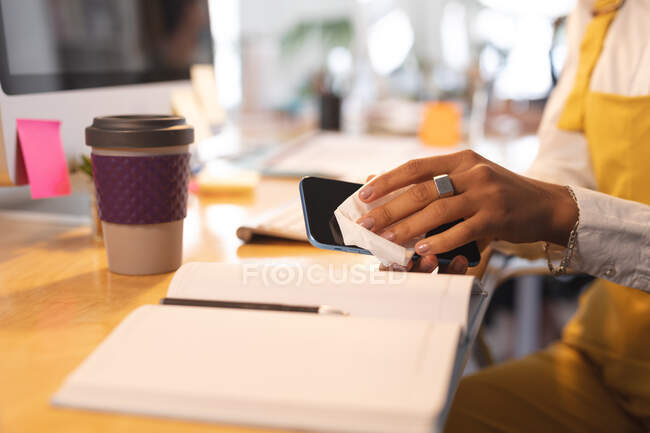 Творческая женщина, сидя за столом в офисе, дезинфицирует свой смартфон. Здоровье и гиперактивность на рабочем месте во время коронавируса Ковид 19 пандемии. — стоковое фото