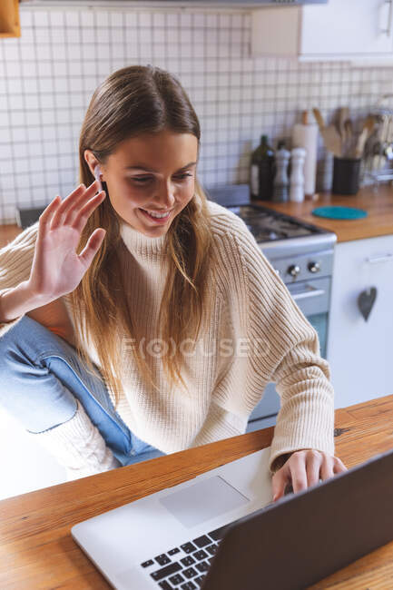 Кавказка проводит время дома, сидит на кухне с ноутбуком с наушниками, улыбается и машет во время видеочата. Социальное дистанцирование во время изоляции коронавируса Covid 19. — стоковое фото