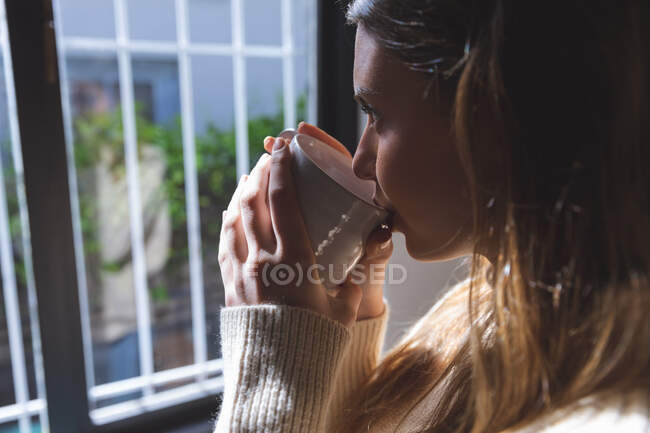 Mujer caucásica pasando tiempo en casa, de pie junto a la ventana, bebiendo de taza verde mirando por la ventana. Distanciamiento social durante el bloqueo de cuarentena del Coronavirus Covid 19. - foto de stock