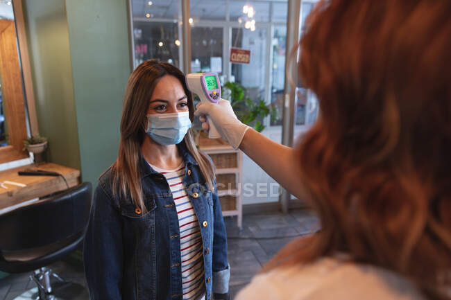 Cabeleireiro feminino caucasiano trabalhando no salão de cabeleireiro, medindo a temperatura de uma cliente caucasiana feminina na máscara facial. Saúde e higiene no local de trabalho durante a pandemia de Coronavirus Covid 19. — Fotografia de Stock