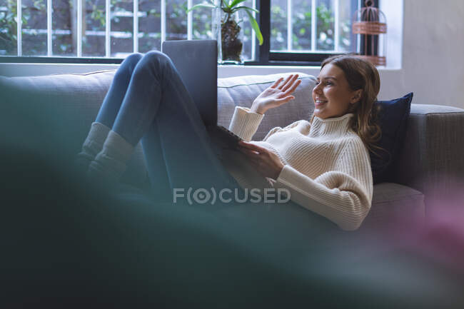 Mujer caucásica feliz pasar tiempo en casa, acostado en el sofá en la sala de estar utilizando el ordenador portátil para videollamada, saludando. Distanciamiento social durante el bloqueo de cuarentena del Coronavirus Covid 19. - foto de stock