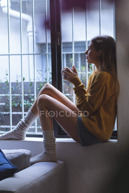 Caucasian woman spending time at home, sitting on windowsill in living room, holding green mug looking out of window. Social distancing during Covid 19 Coronavirus quarantine lockdown. — Stock Photo