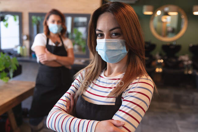 Portrait de femme coiffeuse caucasienne travaillant dans un salon de coiffure portant un masque facial, posant pour une photo avec les bras croisés. Santé et hygiène sur le lieu de travail pendant la pandémie de Coronavirus Covid 19. — Photo de stock
