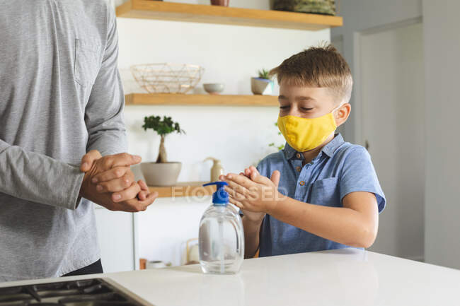 Uomo caucasico a casa con suo figlio, in cucina, ragazzo con la maschera facciale, che si disinfetta le mani. Distanza sociale durante il blocco di quarantena Covid 19 Coronavirus. — Foto stock