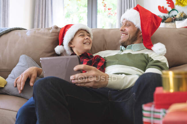 Uomo caucasico a casa con suo figlio a Natale, indossando cappelli di Babbo Natale seduto in soggiorno utilizzando tablet digitale. Distanza sociale durante il blocco di quarantena Covid 19 Coronavirus. — Foto stock