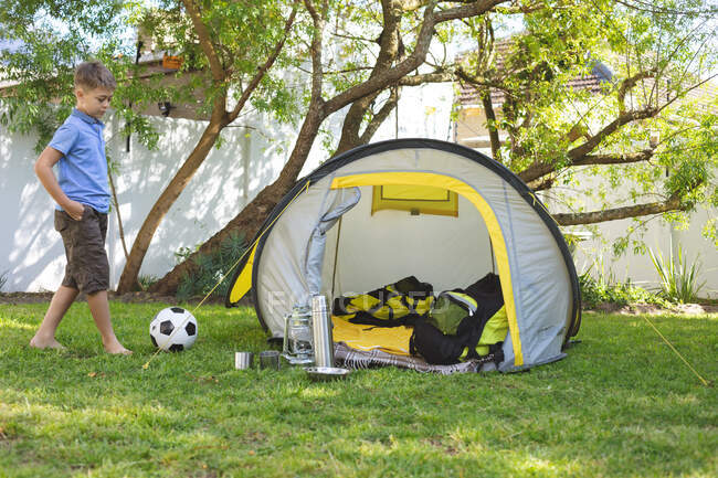 Кавказский мальчик проводит время в саду летом, гуляя с футболом рядом с палаткой под солнцем. Социальное дистанцирование во время изоляции коронавируса Covid 19. — стоковое фото