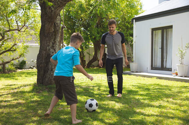 Caucasian man spending time with his son together, playing football in garden. Social distancing during Covid 19 Coronavirus quarantine lockdown. — Stock Photo