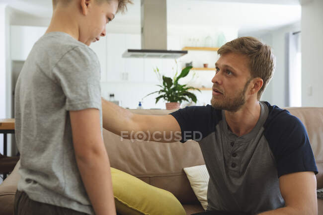 Белый мужчина дома с сыном вместе, сидит на диване в гостиной, отец смотрит на грустного мальчика. Социальное дистанцирование во время изоляции коронавируса Covid 19. — стоковое фото