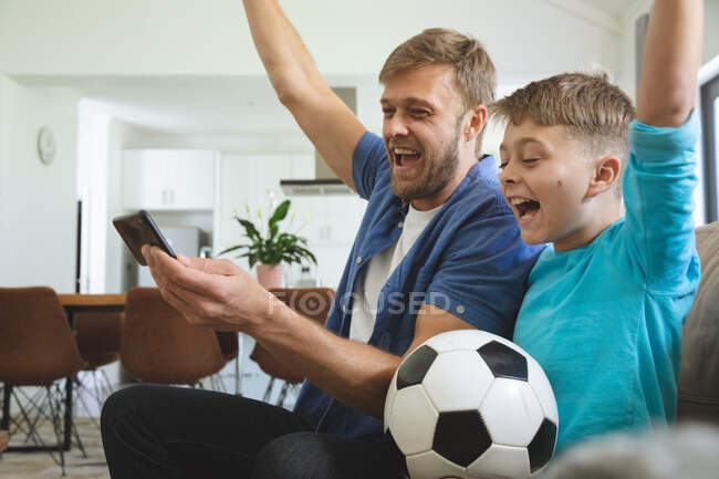 Kaukasischer Mann zu Hause mit seinem Sohn zusammen, auf dem Sofa im Wohnzimmer sitzend, Fußballspiel auf dem Smartphone guckend, jubelnd. Soziale Distanzierung während Covid 19 Coronavirus Quarantäne Lockdown. — Stockfoto