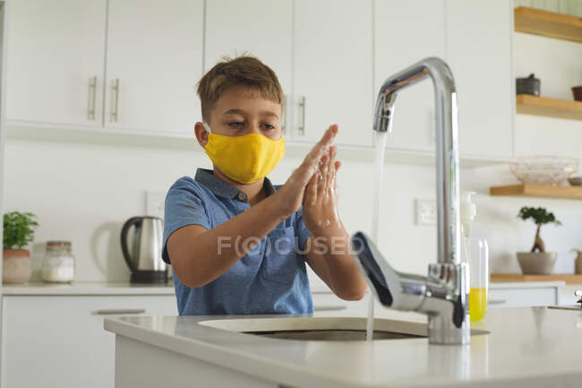 Kaukasischer Junge, der sich zu Hause in der Küche die Hände wäscht und eine gelbe Gesichtsmaske trägt. Soziale Distanzierung während Covid 19 Coronavirus Quarantäne Lockdown. — Stockfoto
