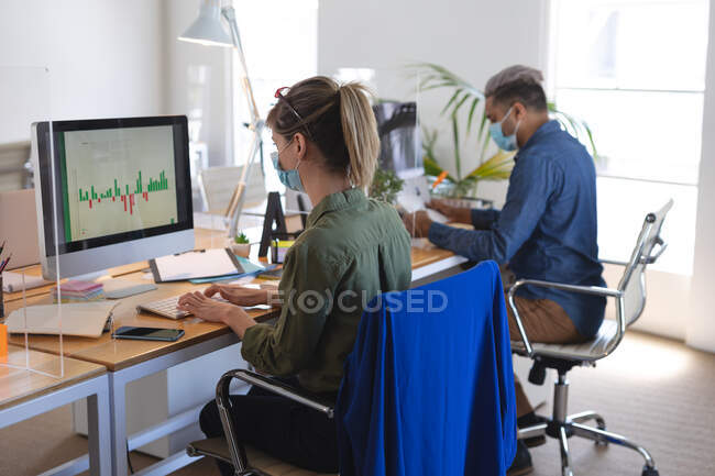 Kaukasische Frau, die mit ihrem Kollegen am Schreibtisch in einem modernen Büro sitzt, Gesichtsmaske trägt und einen Computer benutzt. Gesundheit und Hygiene am Arbeitsplatz während der Coronavirus Covid 19 Pandemie. — Stockfoto