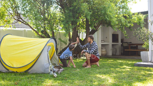 Caucasian man spending time with his son together, camping in garden, sitting by tent, high fiving. Social distancing during Covid 19 Coronavirus quarantine lockdown. — Stock Photo